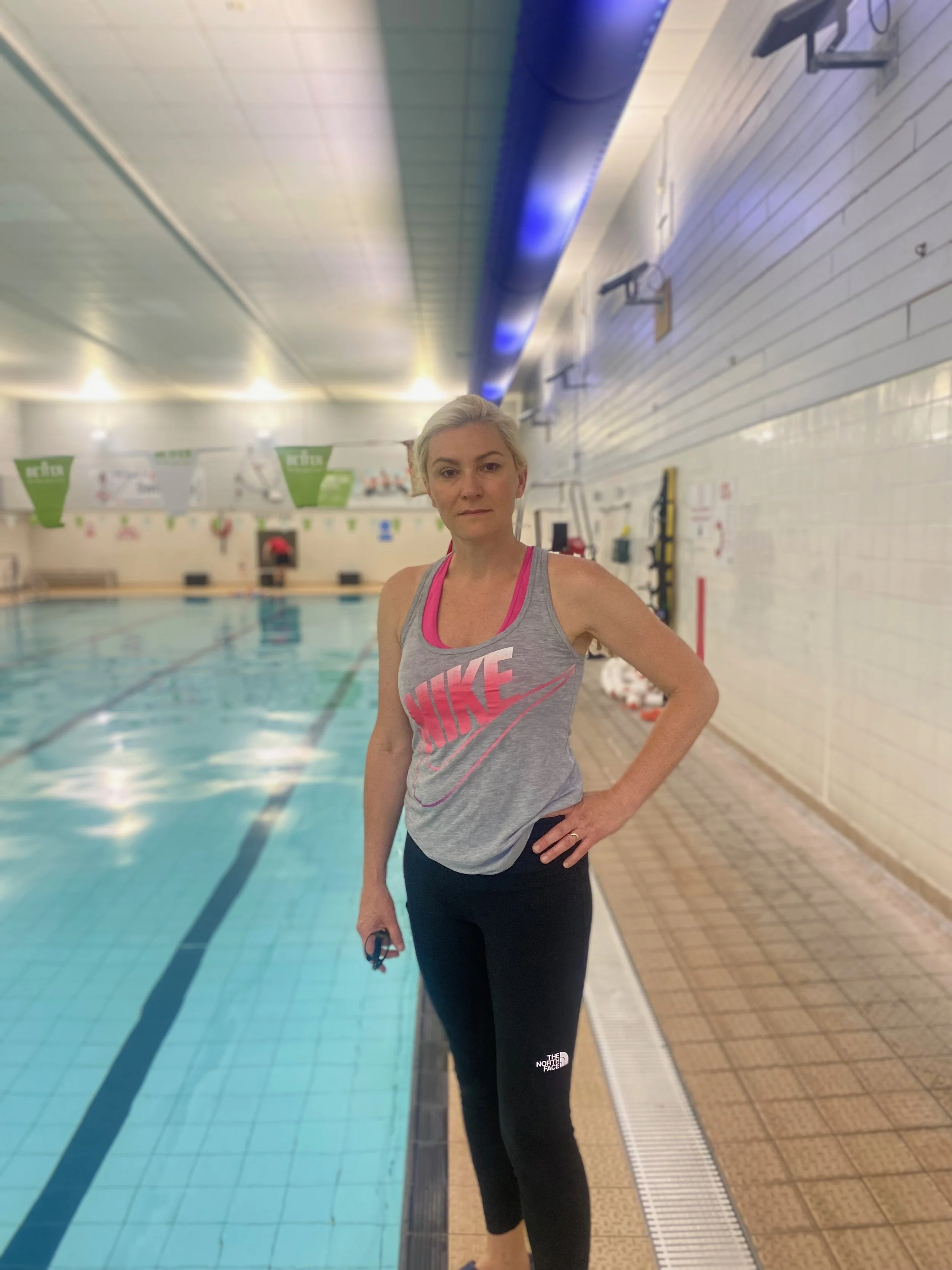 A woman standing by a swimming pool in an indoor facility, wearing a gray Nike tank top and black The North Face leggings, holding goggles in her right hand.