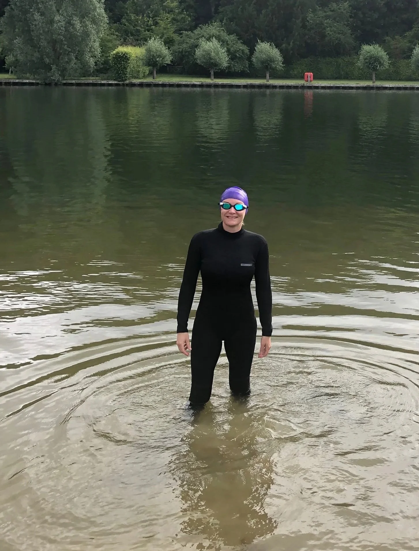 A person standing in a body of water near a riverbank with trees in the background, wearing a black wetsuit, purple swim cap, and mirrored sunglasses, smiling at the camera.