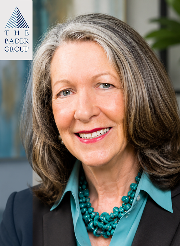 A middle-aged woman with shoulder-length gray hair, smiling and wearing a teal shirt with a matching beaded necklace. She is dressed in a black blazer and is in an indoor setting with modern decor. The top left corner has a logo with a blue triangle and the text "The Bader Group."