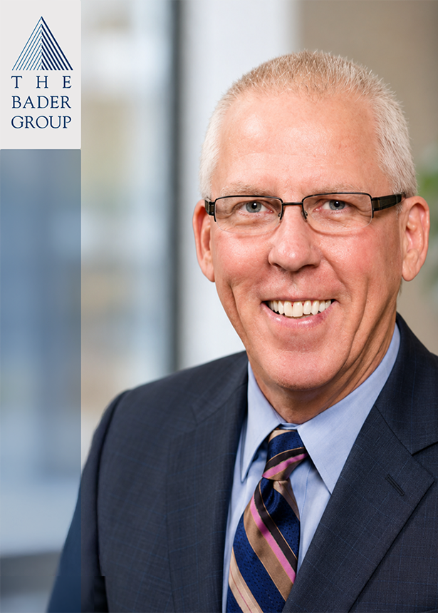 A smiling man wearing glasses, a dark suit, and a striped tie in an office setting, with a company logo for The Bader Group visible on the left side of the image.