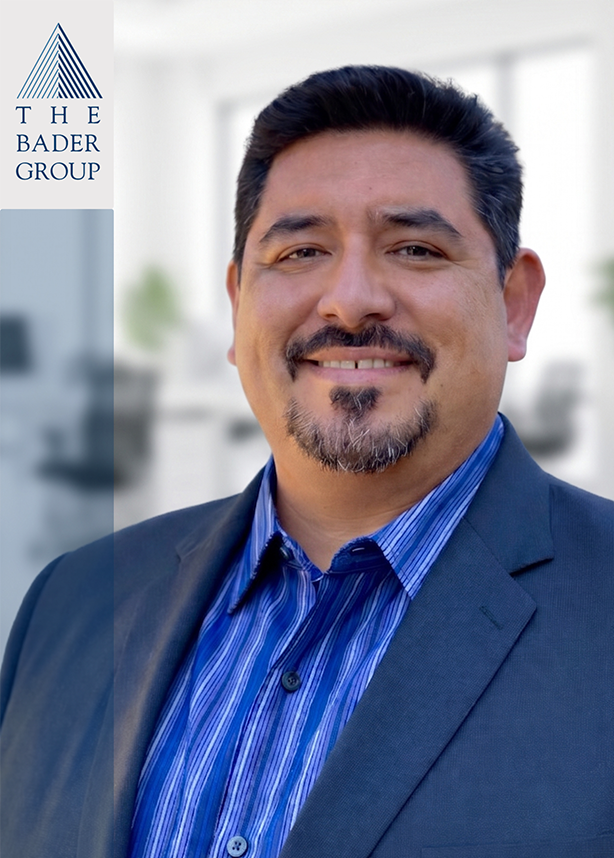 Professional headshot of a man with dark hair and beard, wearing a suit and blue striped shirt, smiling in an office setting with the Bader Group logo on the left.