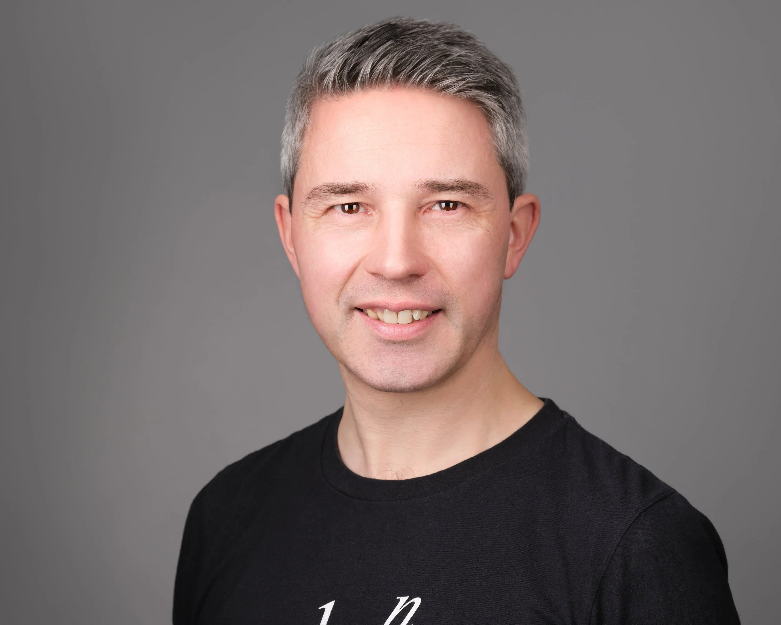 A man with short, gray styled hair, light skin, smiling, wearing a black shirt, against a gray background.