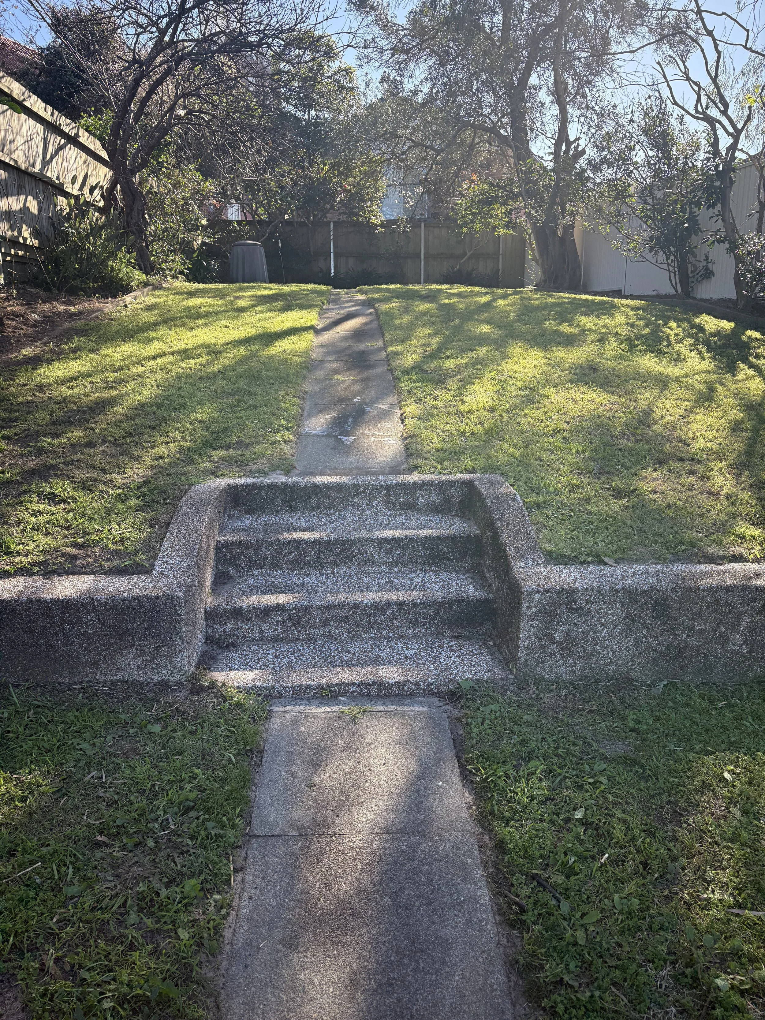 Concrete stairs leading to a sidewalk in a backyard with a grassy lawn, trees, and a wooden fence.
