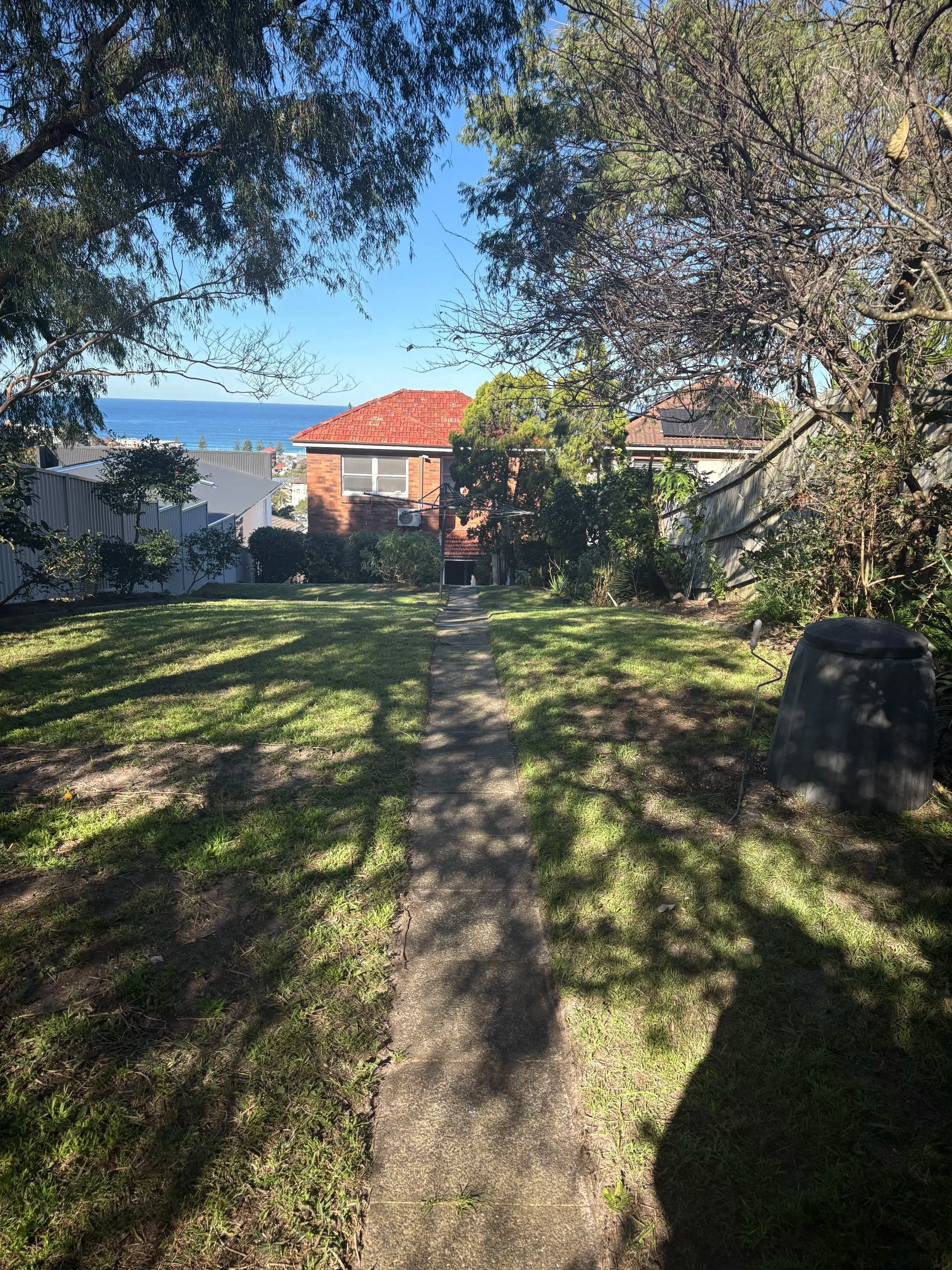 A sidewalk running through a grassy backyard with trees and bushes on both sides. Houses are visible in the background, with a view of the ocean and blue sky beyond.