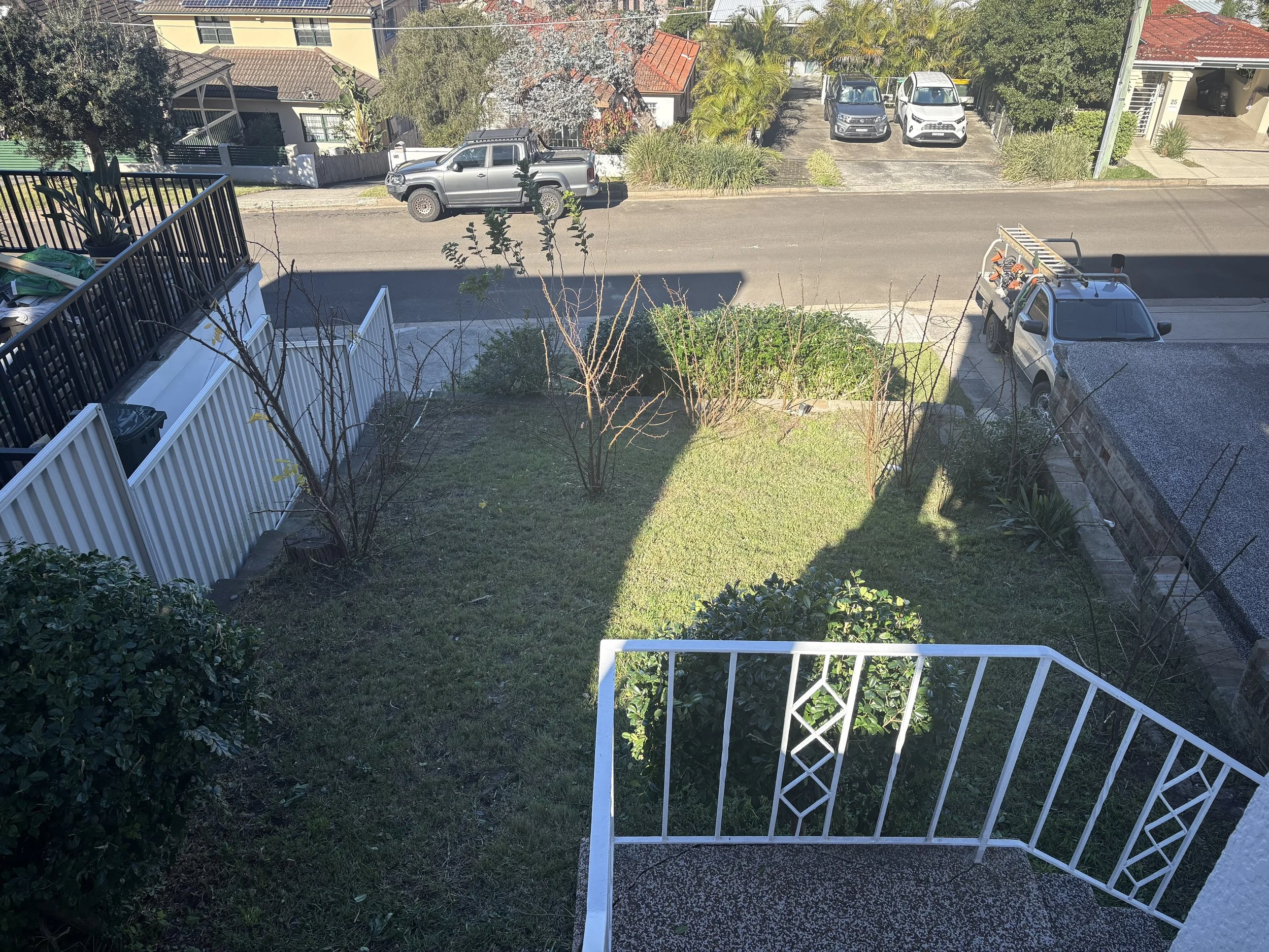 View from a balcony showing a backyard with a grassy area, shrubbery, and a white fence. Beyond the fence, a street with parked vehicles including a silver truck, a white car, and a gray car is visible. There are trees and neighboring houses in the b