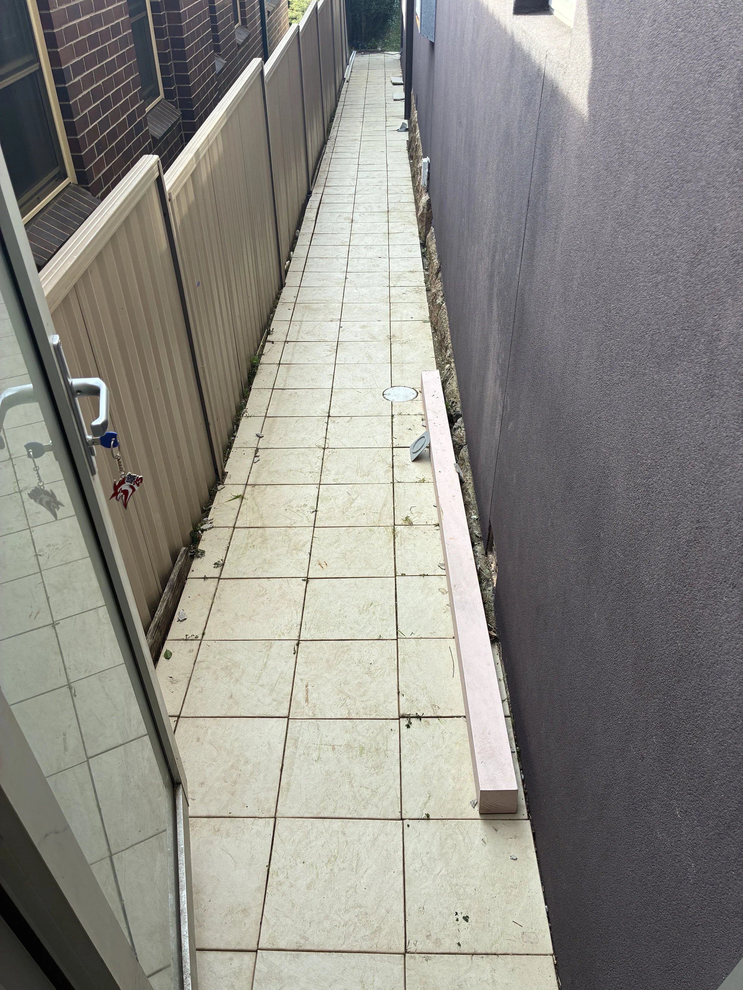 View of a narrow outdoor walkway with beige tiled flooring, bordered by a dark gray wall on the right and a beige fence on the left. The fence has some vertical ridges and is topped with a horizontal metal strip. Several small plants are growing betw