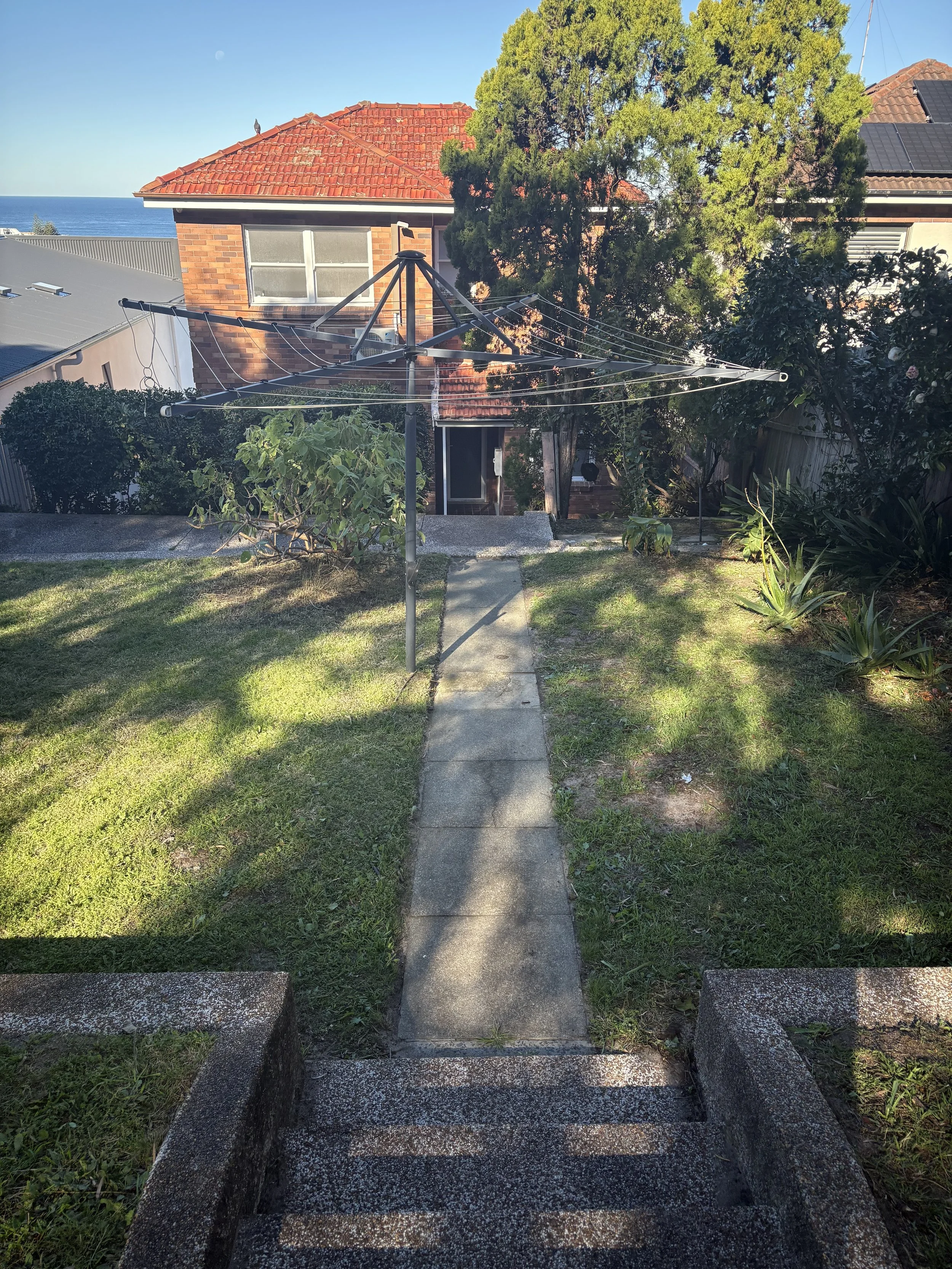 Concrete pathway leading up stairs bordered by grass and shrubs. In the background, a brick house with a red tile roof and trees, with a view of the ocean and a blue sky.