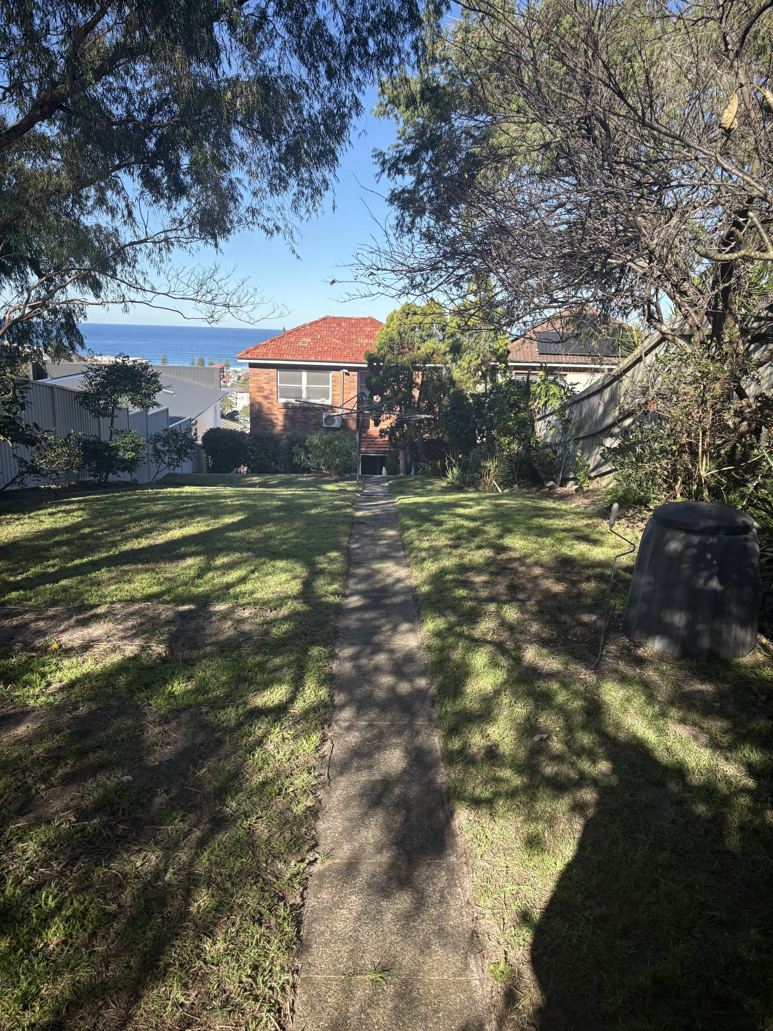 A concrete pathway leading through a grassy backyard, surrounded by trees with leaves and shadows, and residential houses in the background near the coastline with the ocean visible under a clear blue sky.