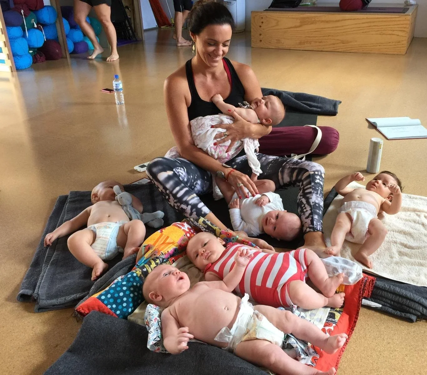 A woman sitting on a mat in a room filled with babies, holding one baby in her arms and surrounded by other babies lying on the mat, some playing with toys. The woman is smiling and dressed in patterned leggings and a black tank top. Babies are in diapers, and a water bottle and notebooks are visible nearby.