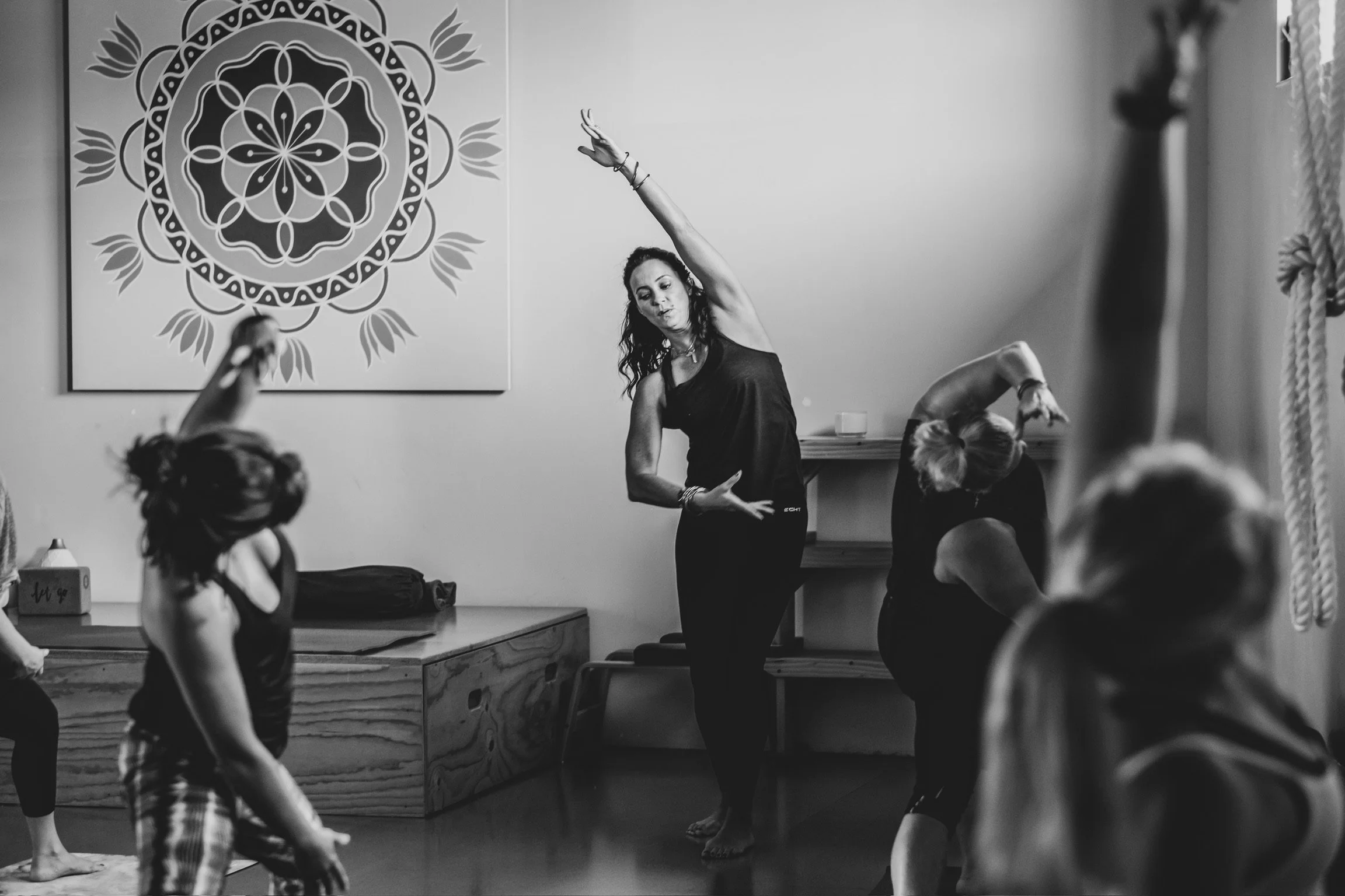 A woman leading a yoga class with children, performing a stretching pose, in a room with a large mandala artwork on the wall.