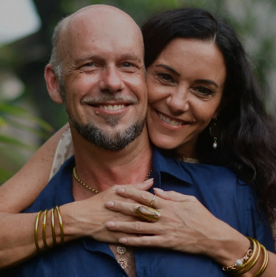 A middle-aged man with a beard and mustache smiling, being embraced from behind by a woman with long dark hair, both smiling, outdoors with blurred greenery in the background.