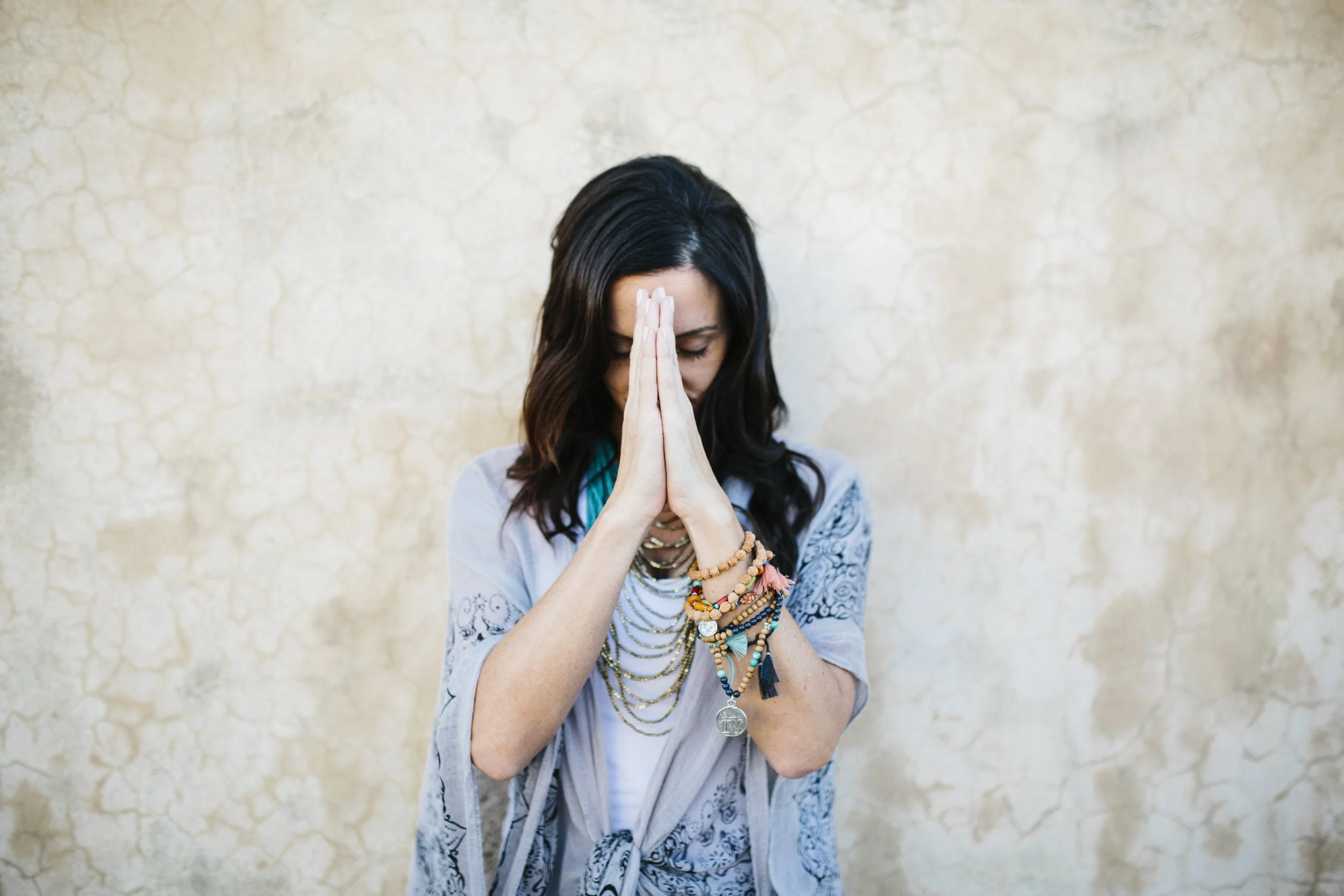 A woman with dark wavy hair, wearing layered necklaces and bracelets, stands with her hands pressed together in prayer or meditation against a textured beige wall.