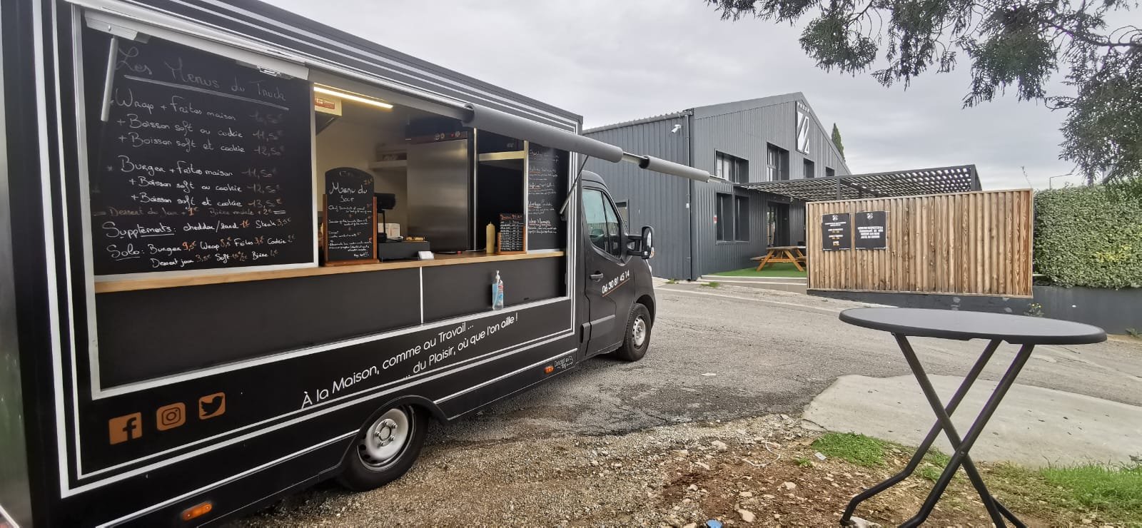 Un camion de nourriture noir stationné à l'extérieur avec un menu écrit à la craie sur un tableau noir, une petite table ronde pliante en face, une zone résidentielle avec une maison en métal gris en arrière-plan, et un ciel nuageux.