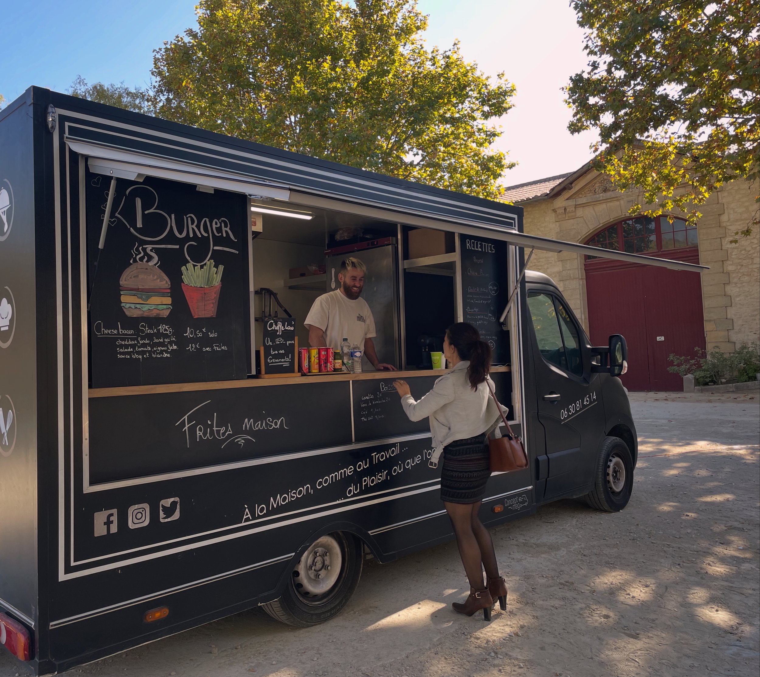 Un camion de restauration de burgers garni de dessins d'un burger et de frites, avec un menu écrit à la craie. Deux personnes discutent devant le camion, dans un cadre extérieur avec un arbre au fond.