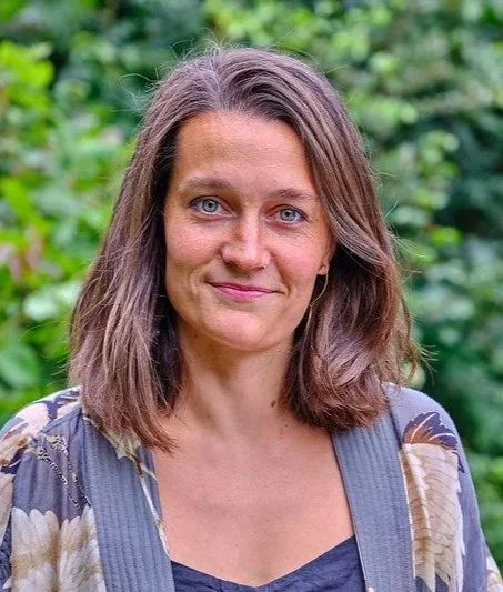 A woman with shoulder-length brown hair and blue eyes outdoors with green foliage background.