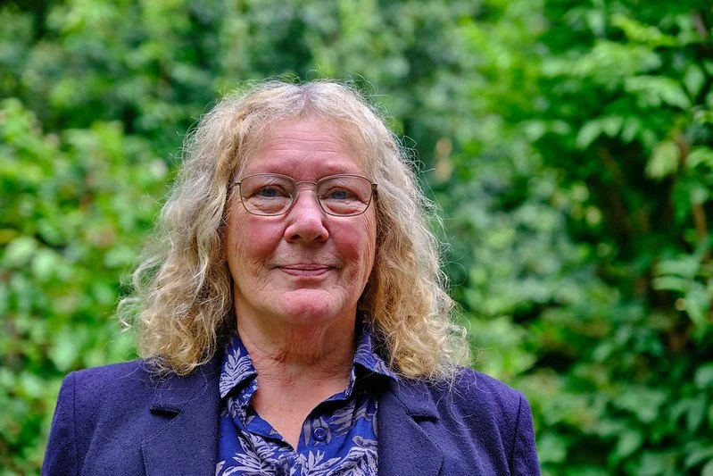 A woman with curly blonde hair and glasses, wearing a dark blazer and patterned shirt, standing outdoors with green foliage in the background.