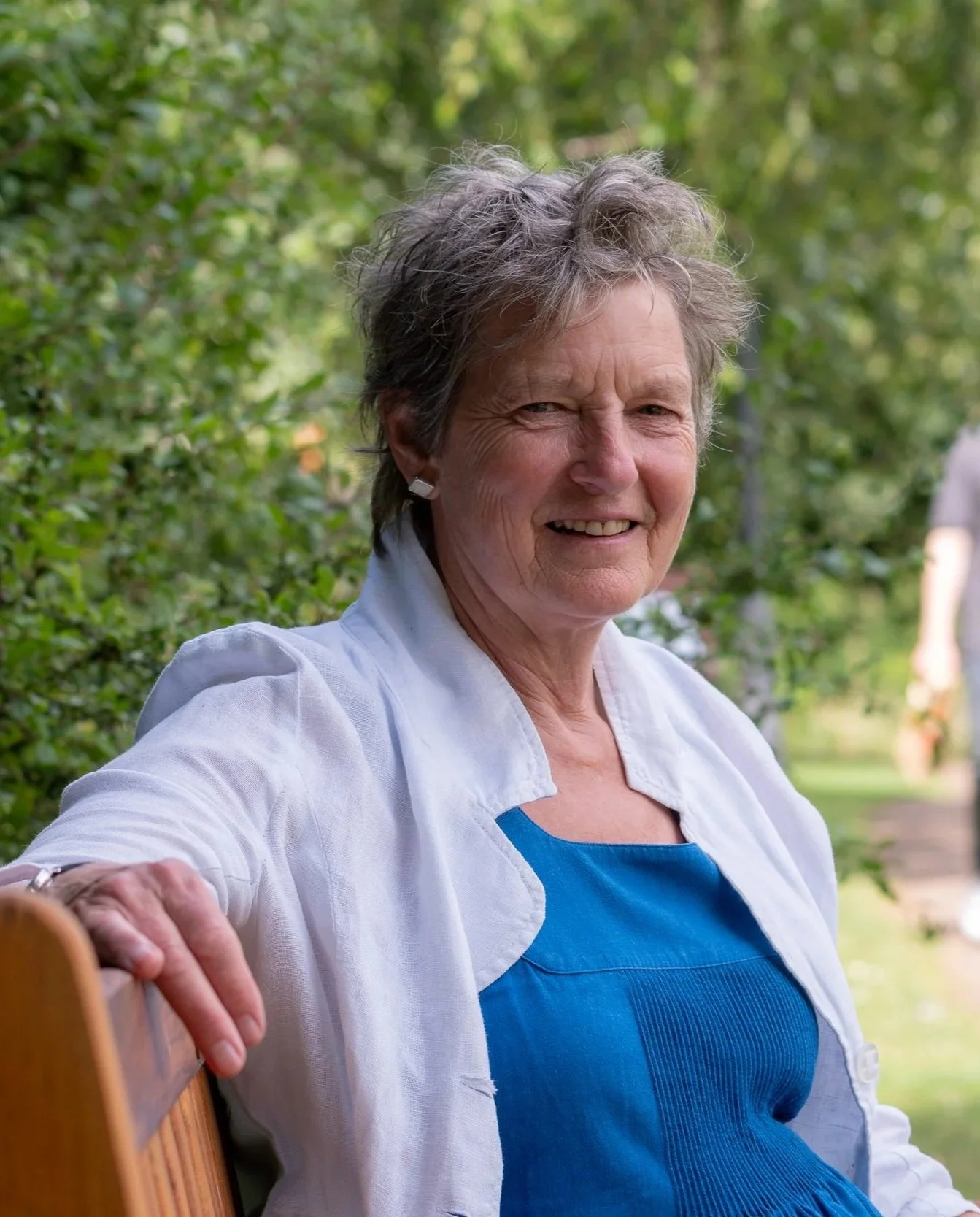A smiling elderly woman with short gray hair sitting outdoors on a wooden bench in a park with green trees in the background, dressed in a blue dress and white cardigan.