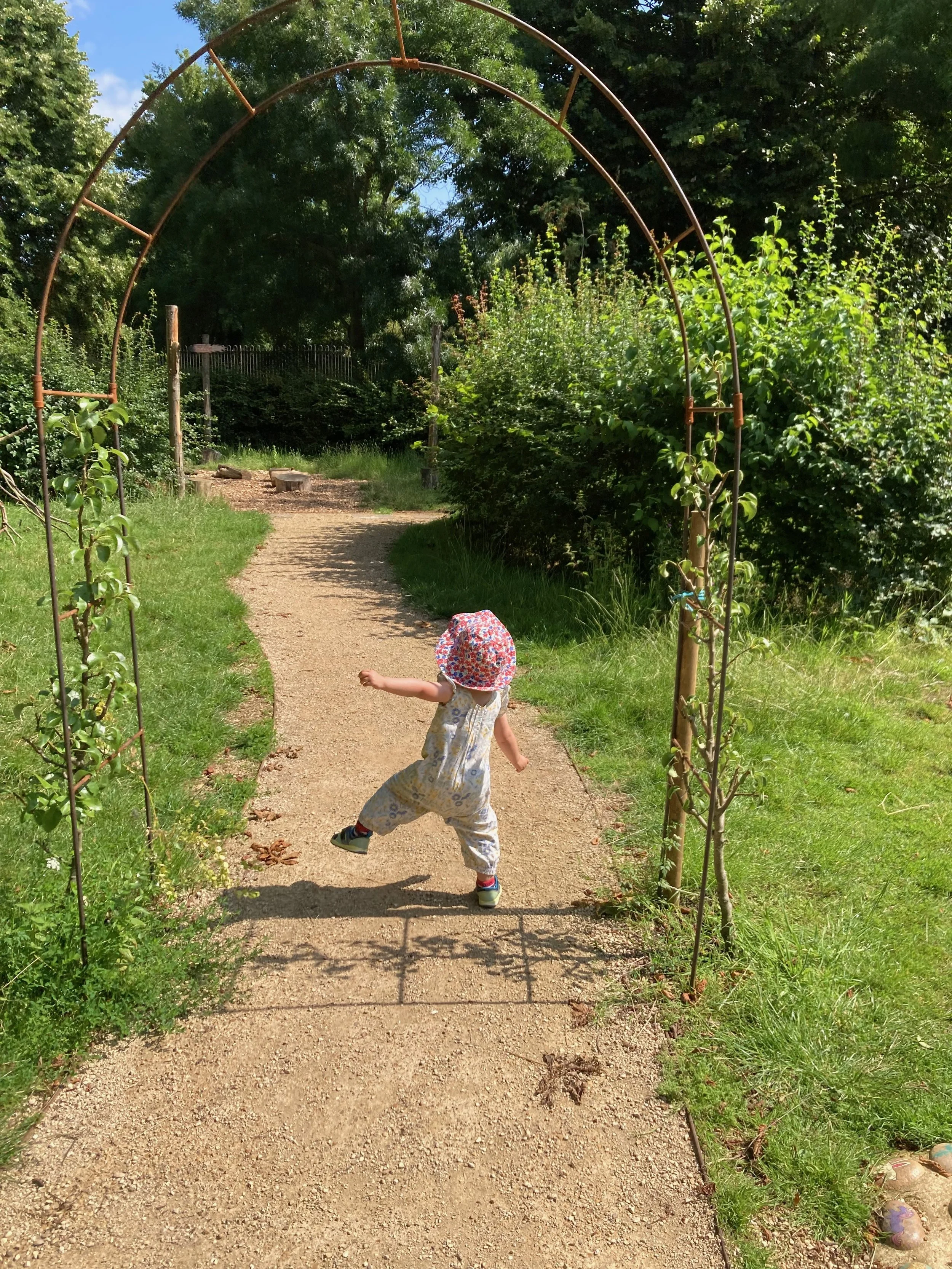 Child jumping in front of a garden arch on a paved path, surrounded by green shrubbery and trees, with a person sitting in the background.