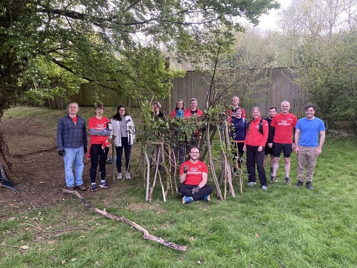 Group of people standing outdoors near a handmade wooden structure decorated with vines and branches, with trees and a wooden fence in the background.