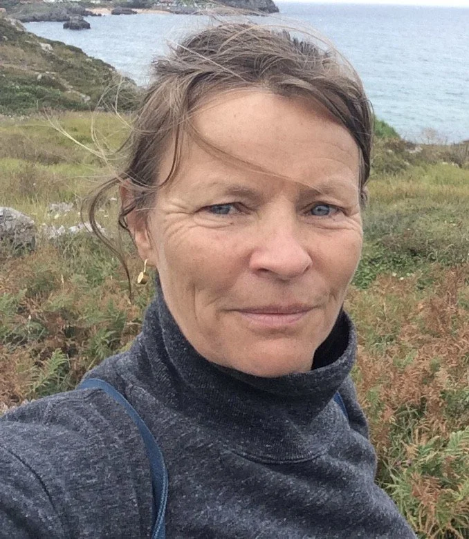 A woman with short light brown hair and blue eyes takes a selfie outdoors in a grassy area near the coast, with the ocean and rocky cliffs visible in the background.