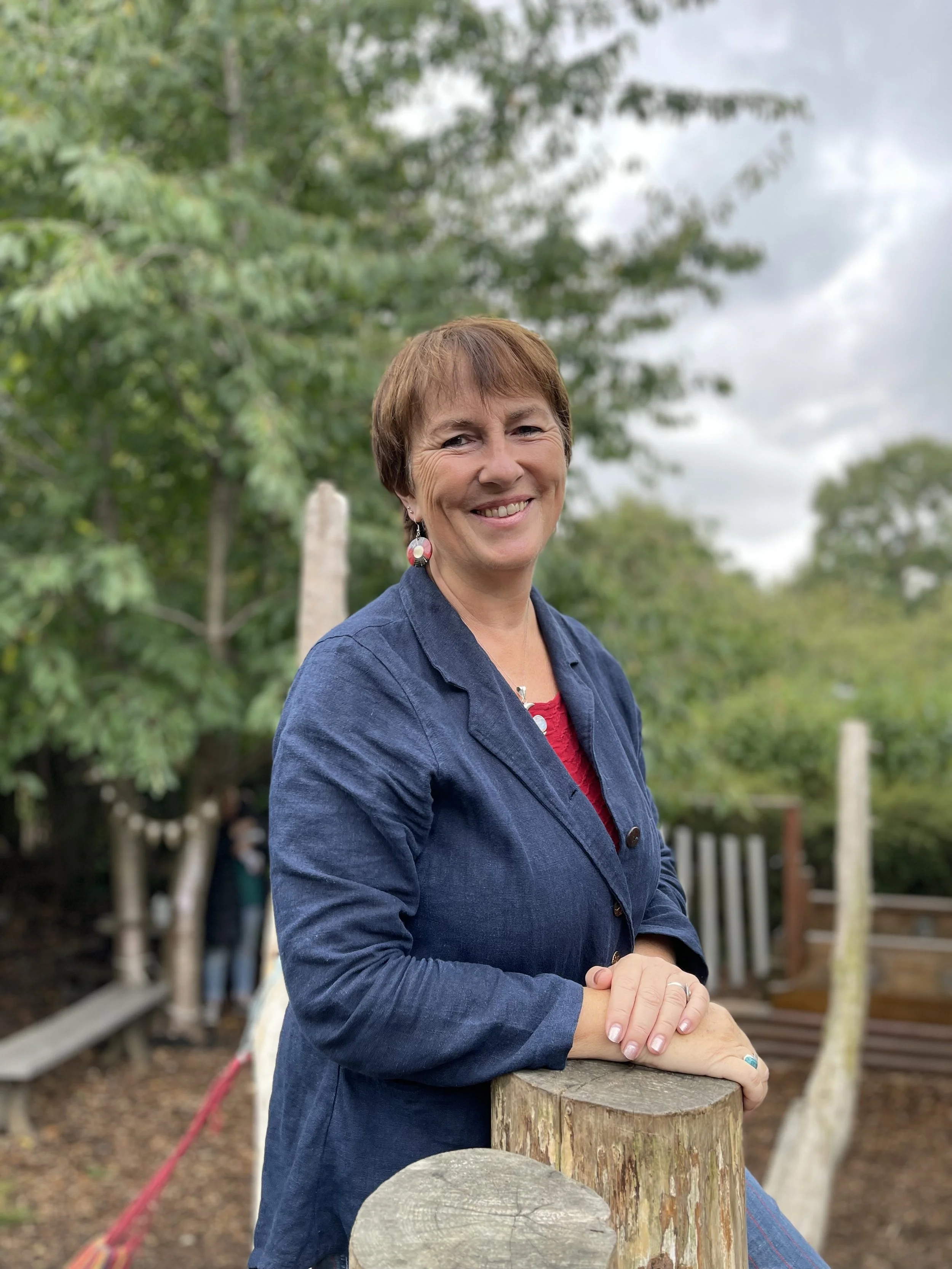 A woman with short brown hair and blue earrings standing outdoors among green foliage.