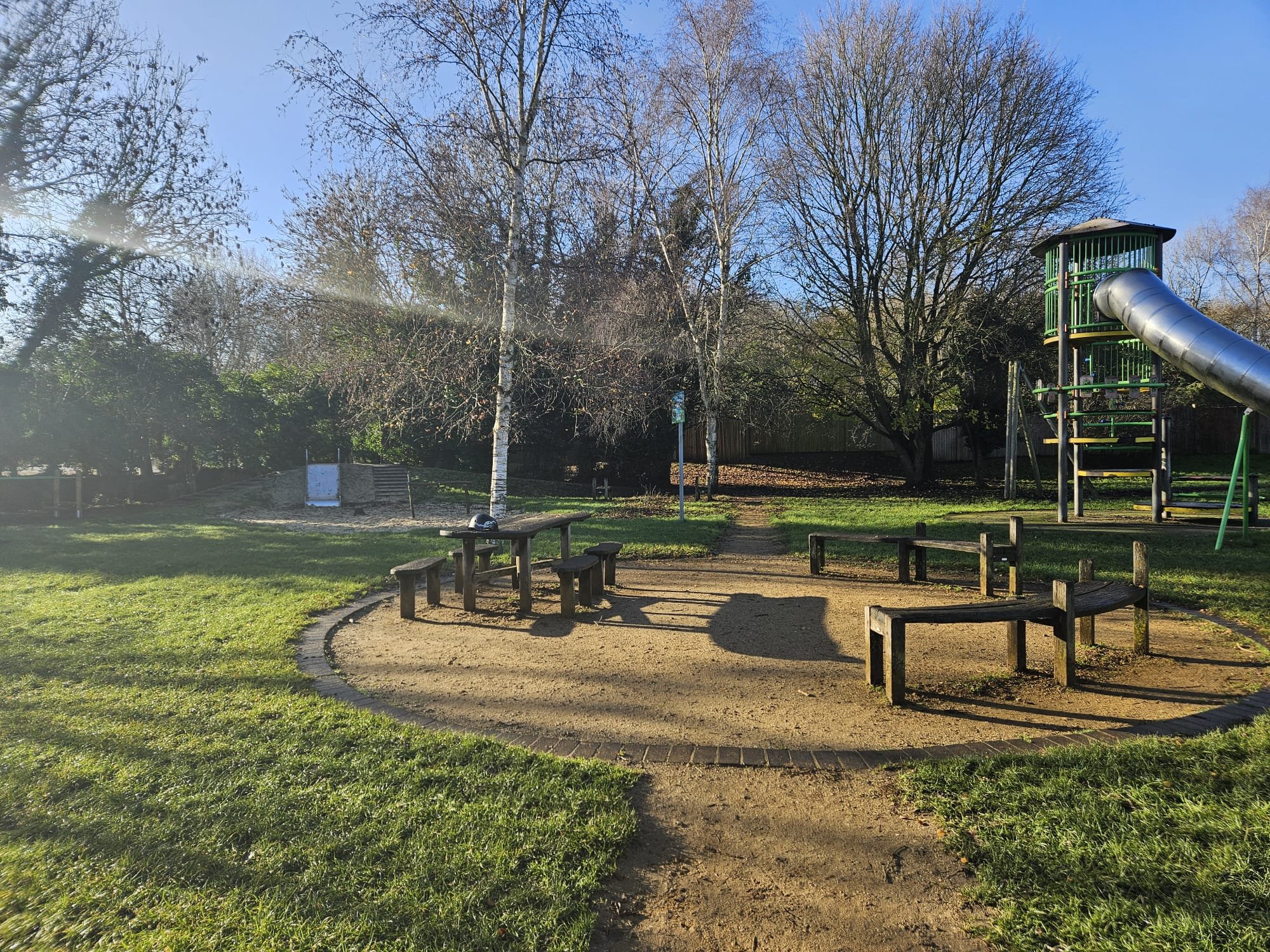 An outdoor playground with a curved dirt pathway, wooden benches, a tall slide, leafless trees, sunny weather, and a blue sky.