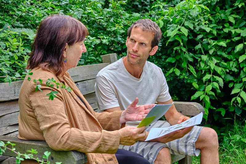 A woman and a man sitting on a park bench, engaged in conversation with documents in their hands, surrounded by green foliage.