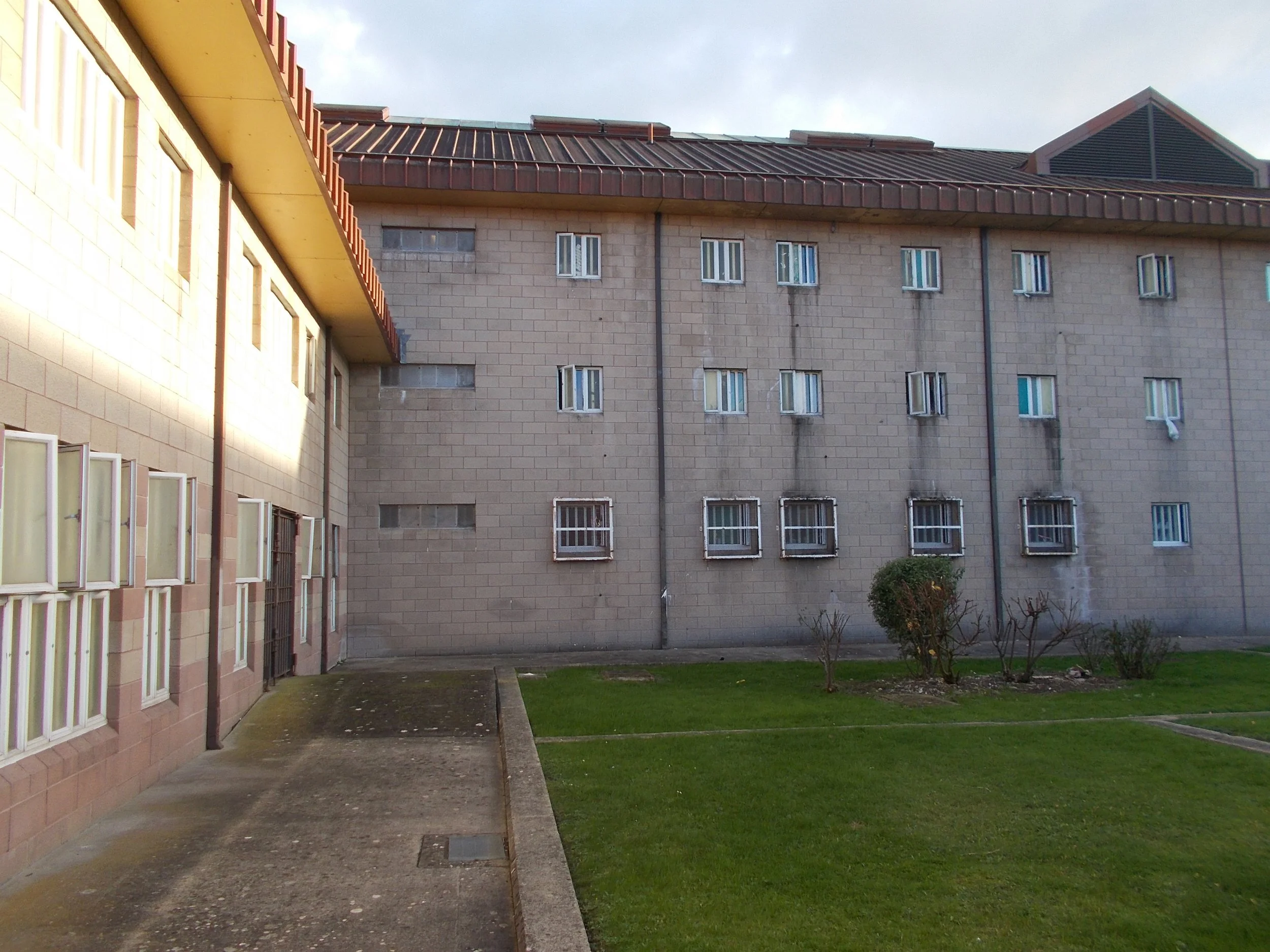 View of a gray multi-story apartment building with small windows, a sloped red roof, and a small courtyard with grass and small trees in the foreground.