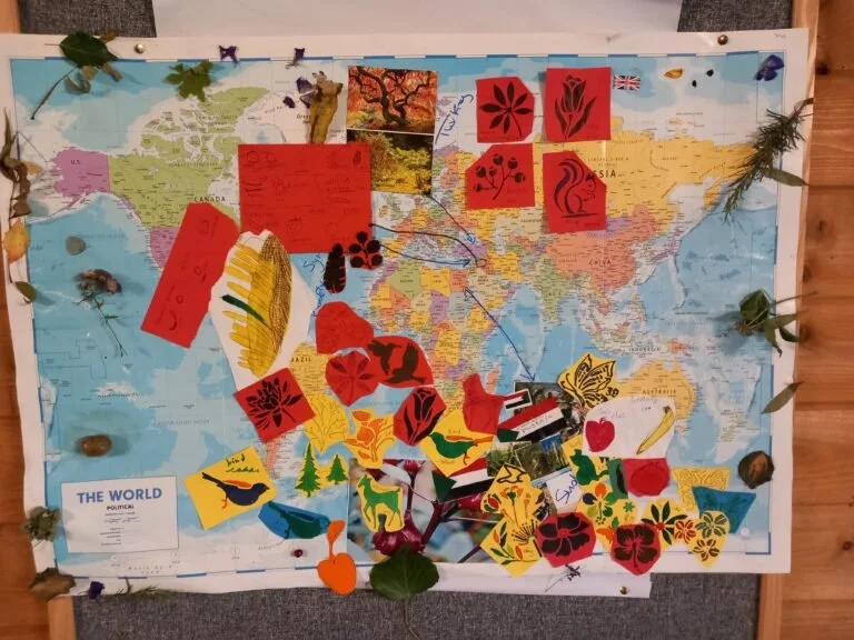 Decorated world map with various colorful paper cutouts of animals, plants, and symbols, along with some photographs and leaves attached.