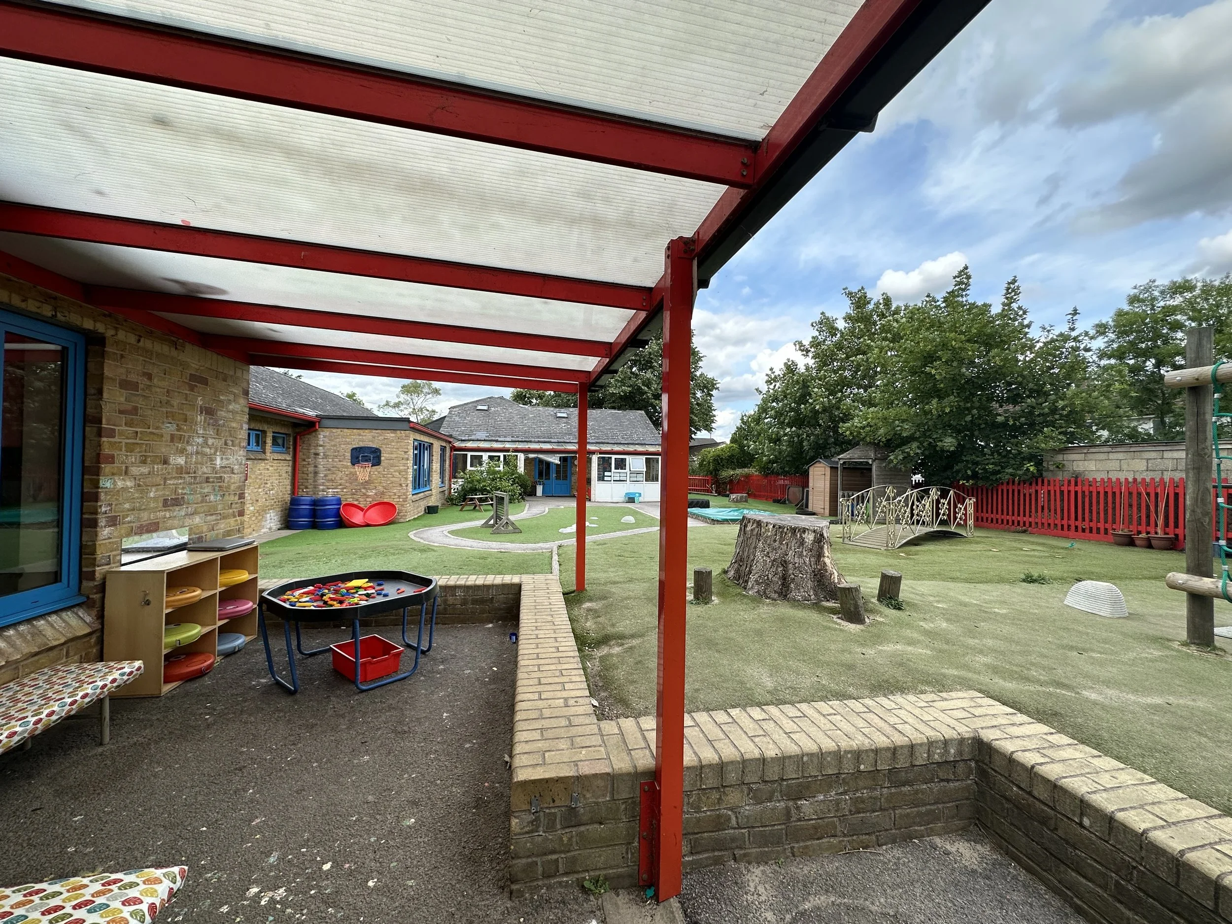 Playground area covered by a translucent roof supported by red beams, with a brick building on the left, a grassy yard with trees, and various playground equipment including tables, a small basketball hoop, and a red coat rack, under a partly cloudy 