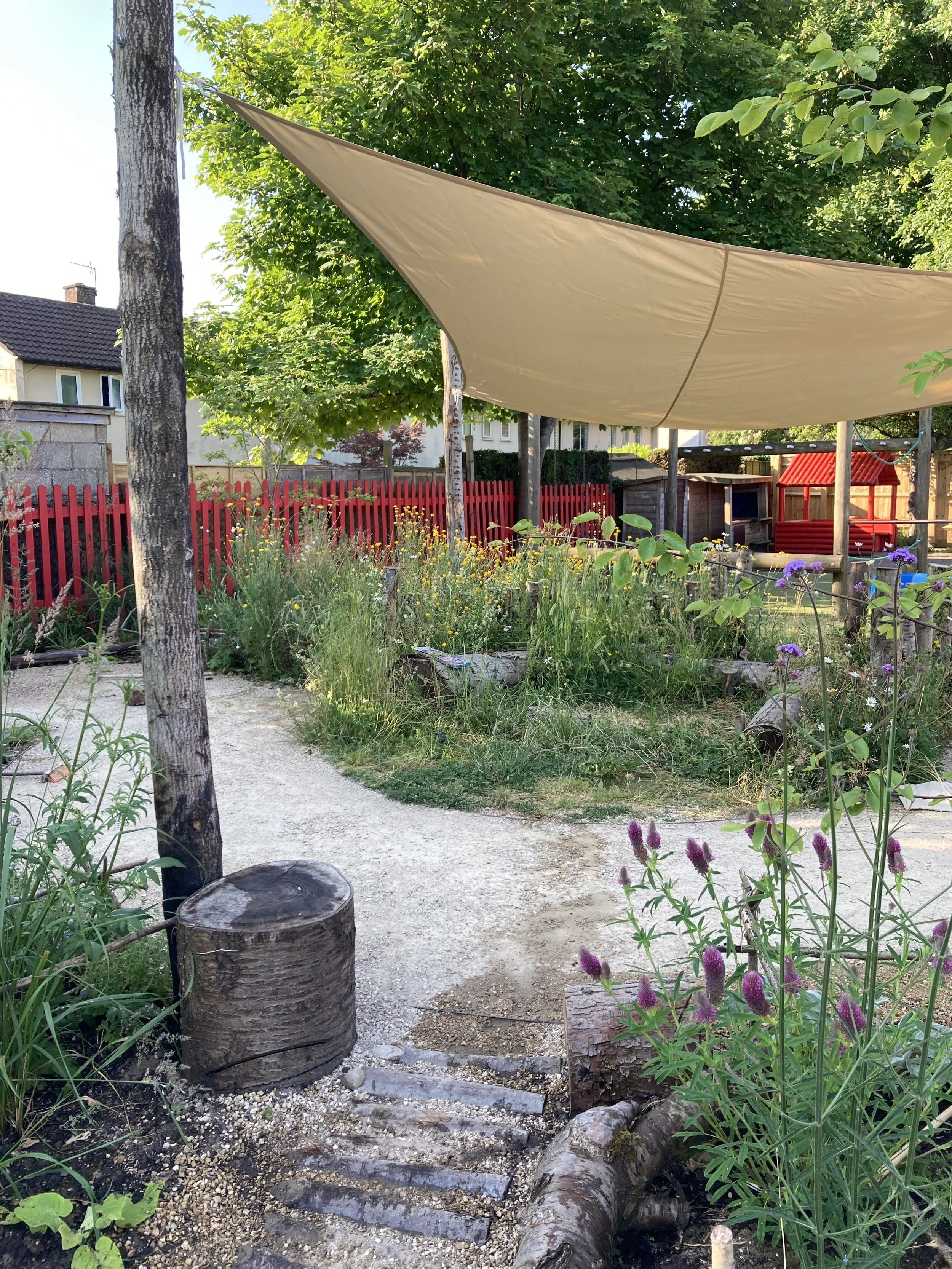 A garden path with wooden steps bordered by plants and flowers, under a large beige shade sail, with trees, a red wooden fence, and a house in the background.