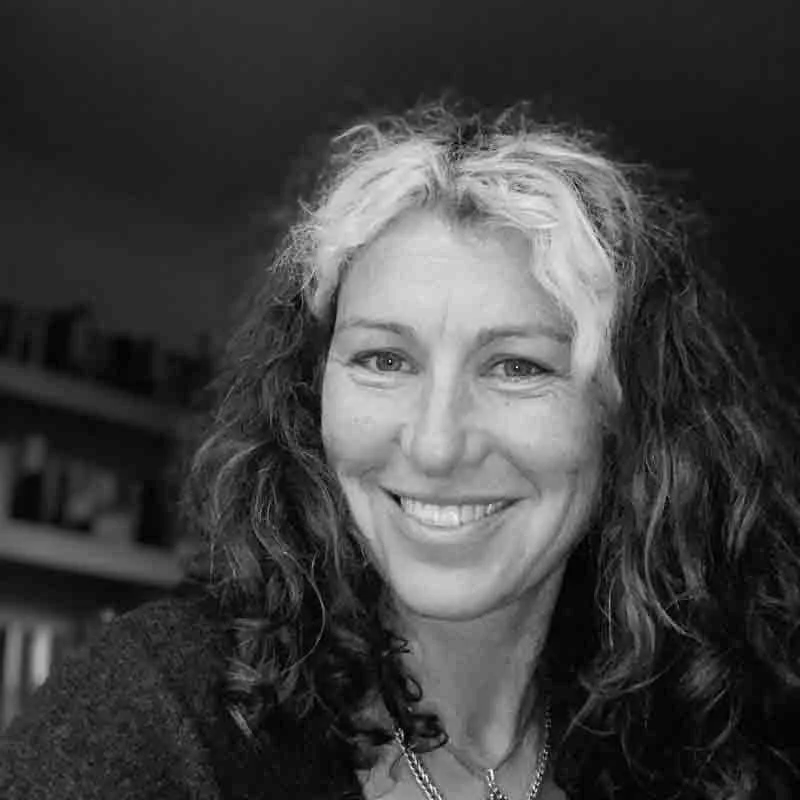 Black and white portrait of a woman with curly hair, smiling, with a bookcase in the background.