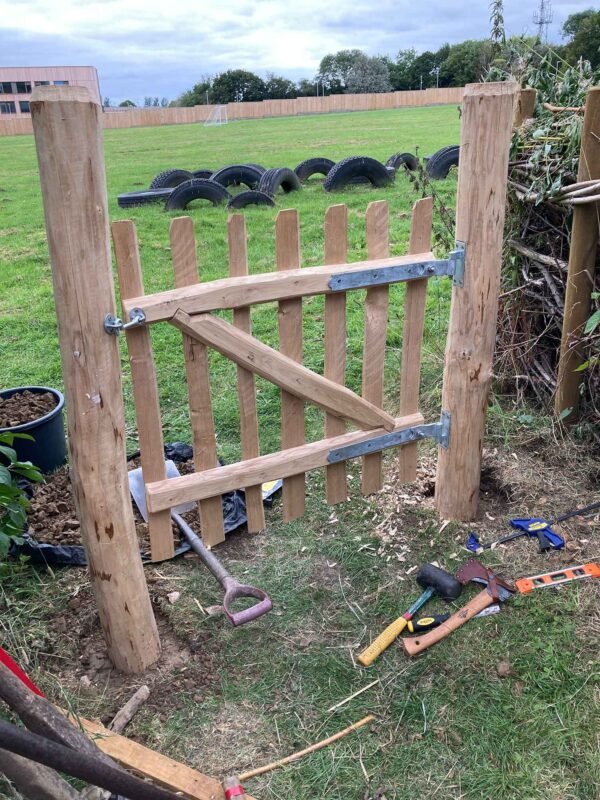 A wooden garden gate under construction with tools like a hammer, saw, and shovel nearby, in a grassy backyard with tire planters and a wooden fence in the background.