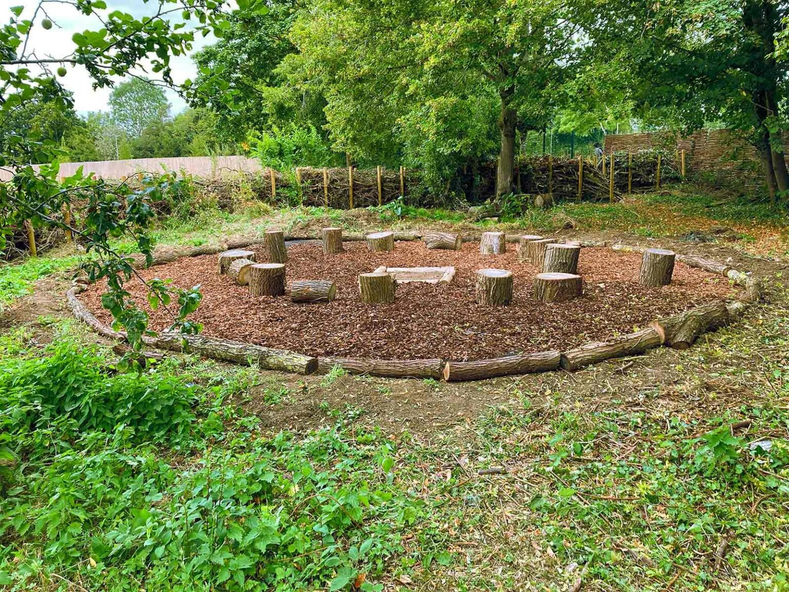 A circular outdoor seating area with wooden logs cut into stumps for seats on a dirt and mulch ground, surrounded by green trees and shrubs, with a simple wooden fence in the background.