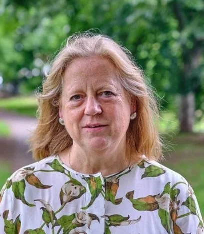 A middle-aged woman with shoulder-length blonde hair, wearing a patterned blouse with green and brown leaf designs, standing outdoors in a park with trees and greenery in the background.