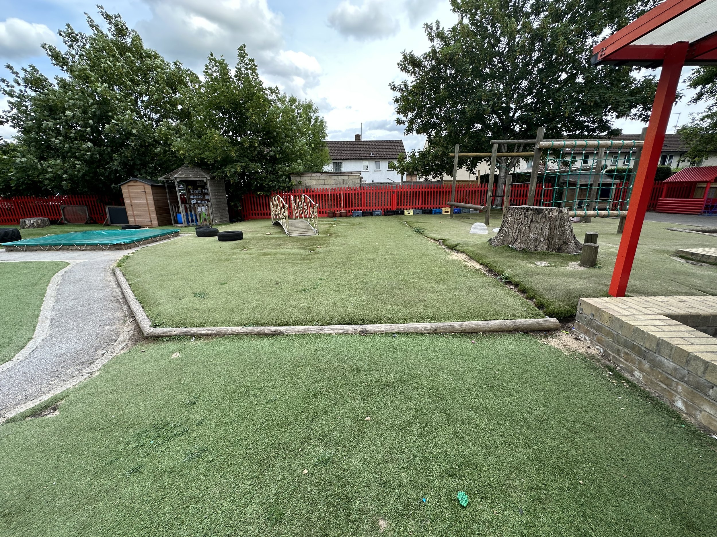 An outdoor playground area with artificial grass, surrounded by a red wooden fence and trees. There is a small bridge, some tires, a garden shed, and climbing structures.