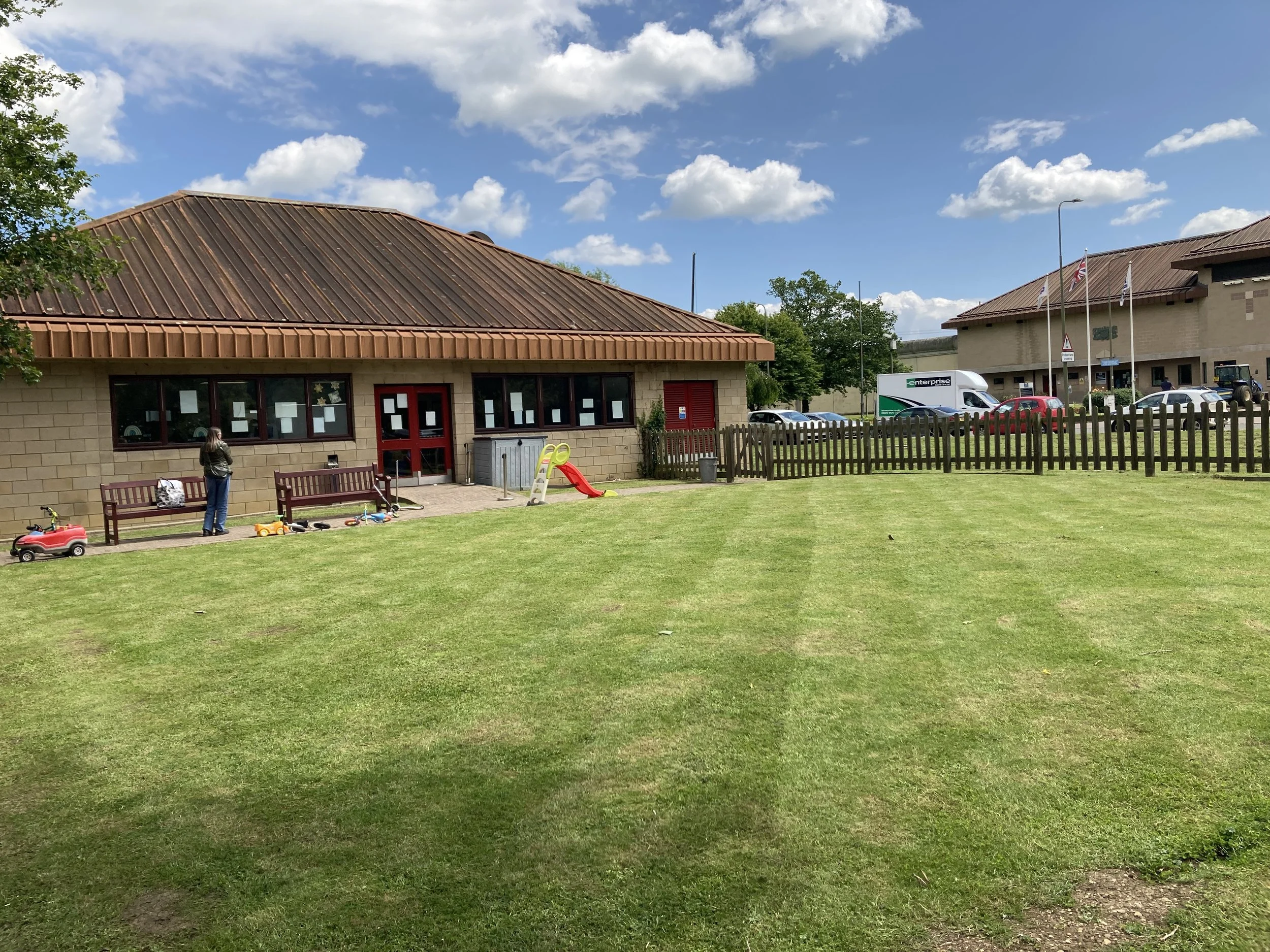 A small, single-story building with beige brick walls, a rust-colored metal roof, and a red door. A woman is standing outside with children's toys scattered on the grass nearby. There are benches in front of the building, a wooden fence, and parked c