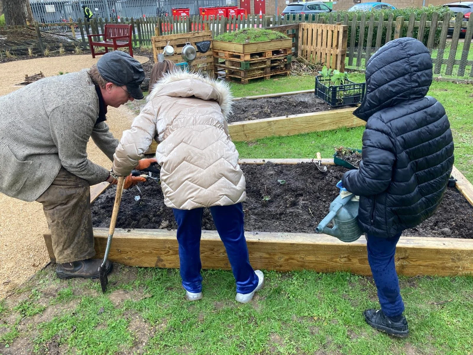 Three people working together planting in a raised garden bed outdoors, with gardening tools and a watering can nearby.