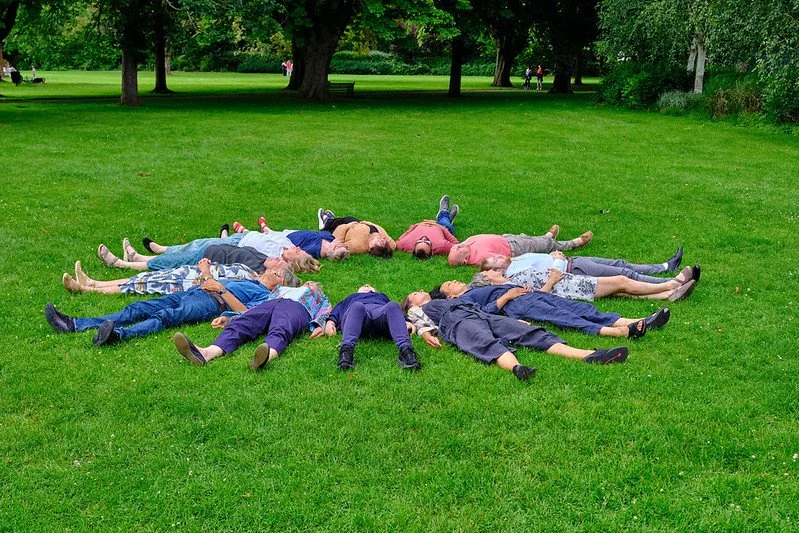 Group of people lying on the grass in a park, arranged in a star shape with heads towards the center and arms and legs extended outward.