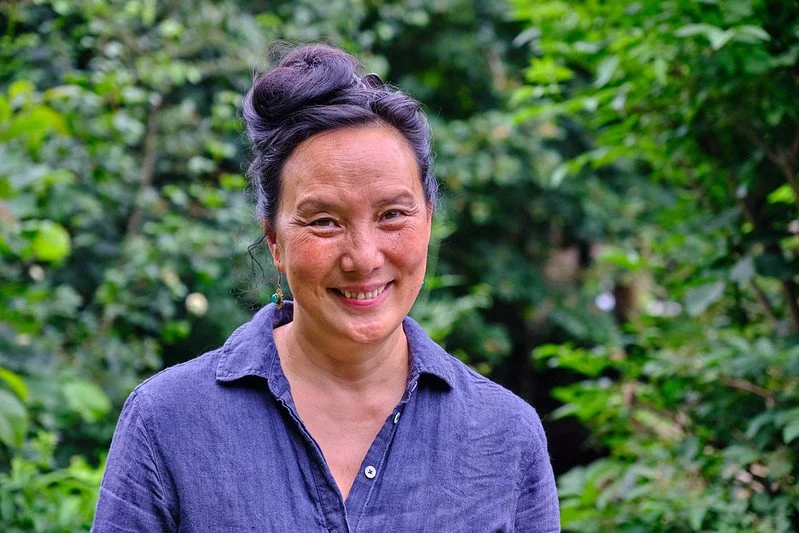 A middle-aged woman with dark purple hair styled in a bun, smiling, wearing a blue button-down shirt, standing outdoors among green trees and foliage.