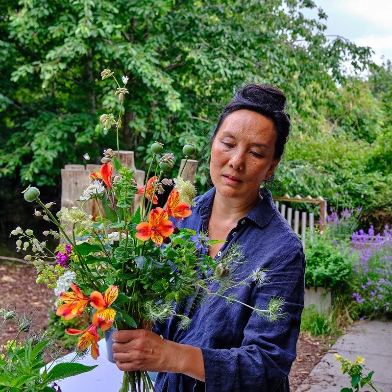 A woman in a blue shirt arranging a colorful bouquet of flowers in a garden with green trees and purple flowers in the background.