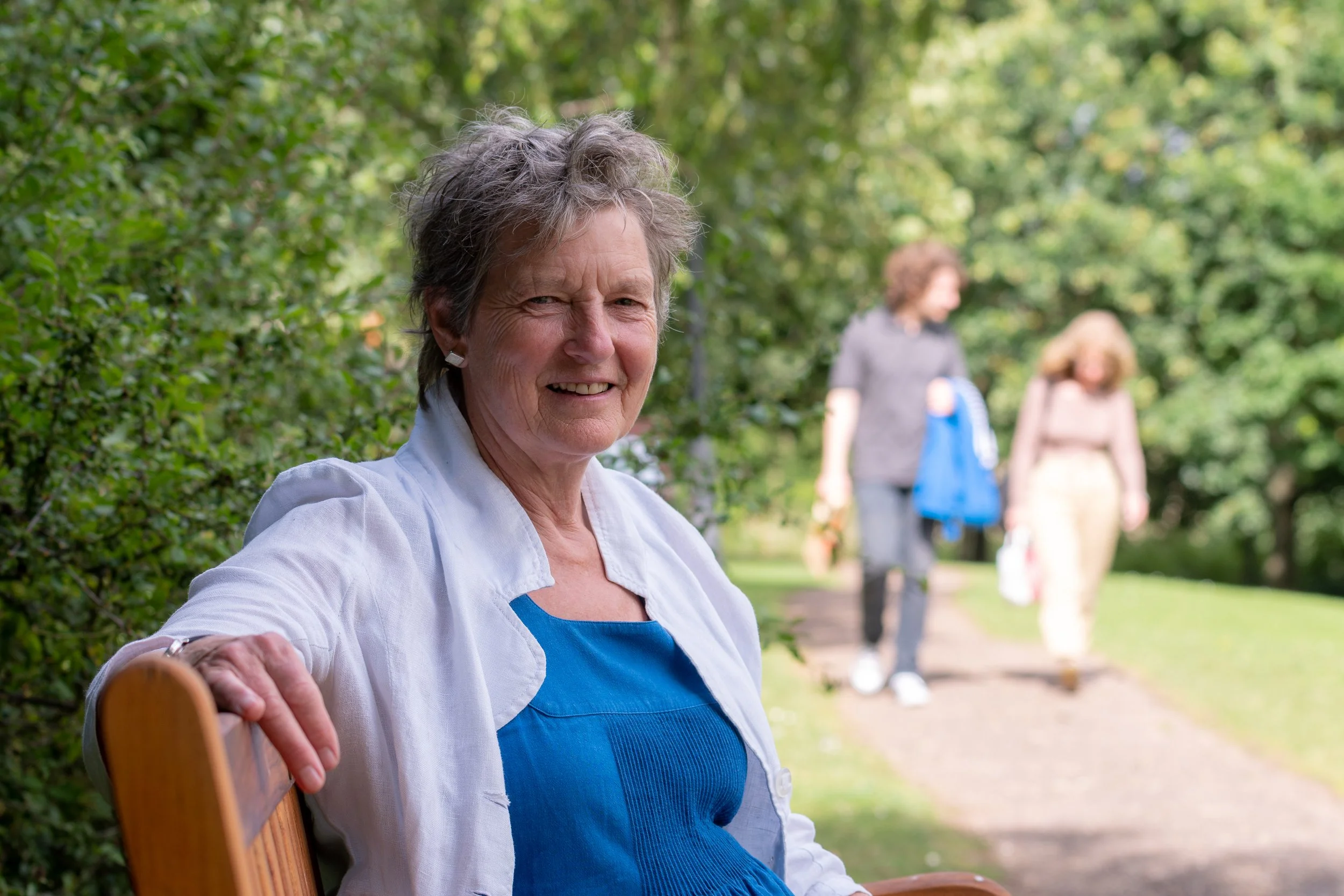 Smiling elderly woman with short gray hair sitting on a wooden bench in a park, wearing a blue dress and white jacket. In the background, two women are walking along a path among green trees.