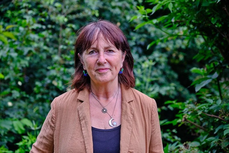 A woman with short brown hair and blue earrings standing outdoors among green foliage.