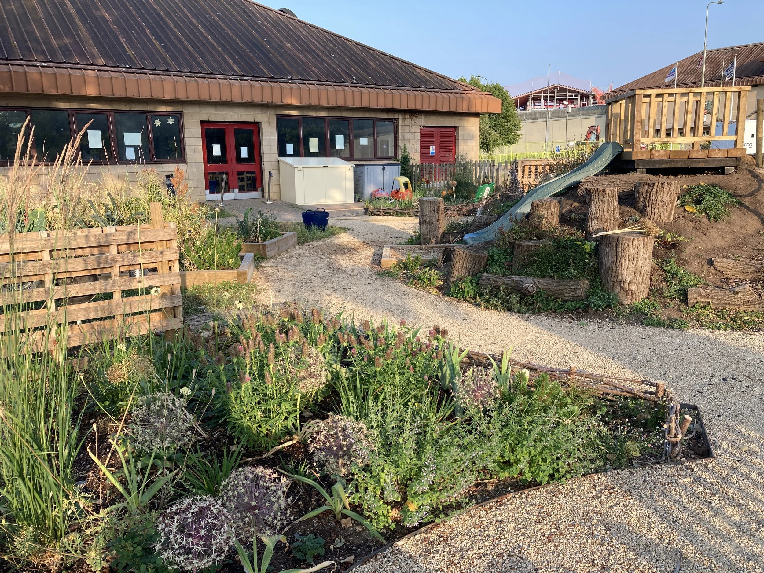 A grassy garden area with plants, flowers, and a small garden fence. There is a building with red doors, large windows, and a metal roof in the background. To the right, there is a small wooden play structure with a slide, and construction work is visible behind a fence.