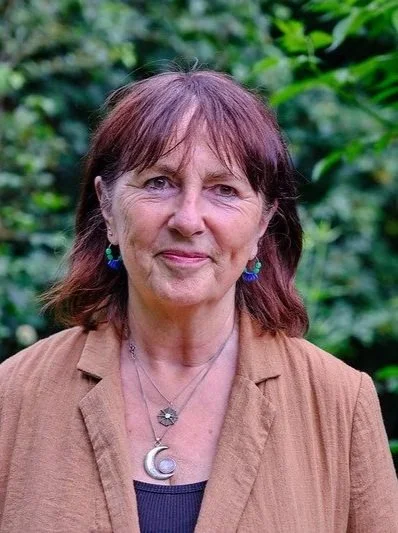 A woman with shoulder-length brown hair, wearing a brown blazer, blue earrings, and layered necklaces, standing outdoors with green foliage in the background.