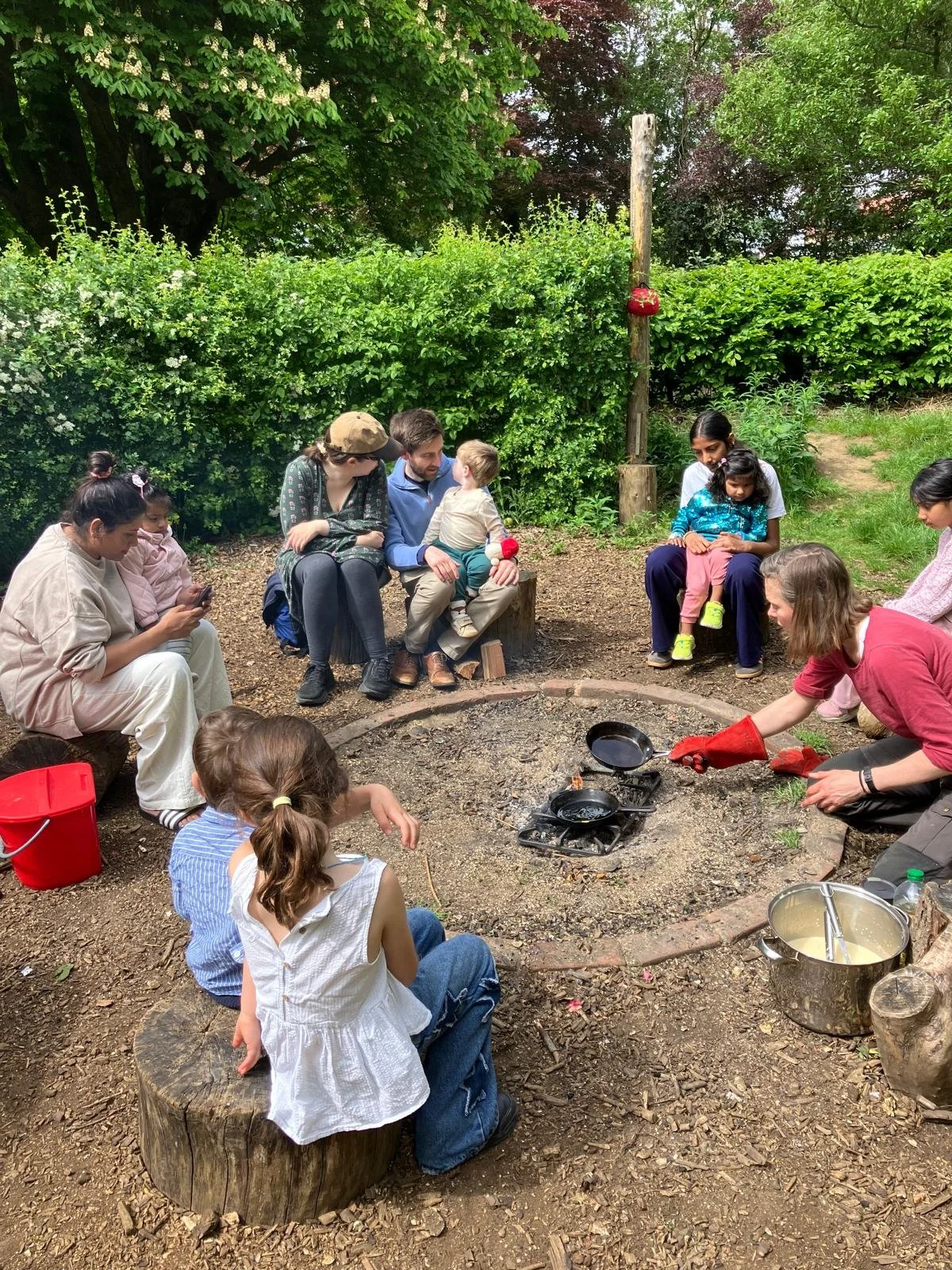 Group of children and adults sitting around a fire pit in a garden, with cooking pans and a woman wearing red oven mitts preparing food over the fire.