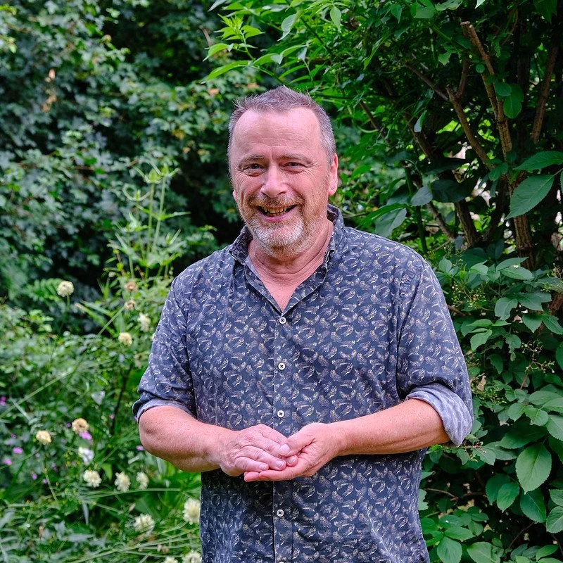 A middle-aged man with short gray hair, a beard, and a mustache, smiling and standing outdoors in a lush garden with green foliage, wearing a patterned button-up shirt with rolled-up sleeves.