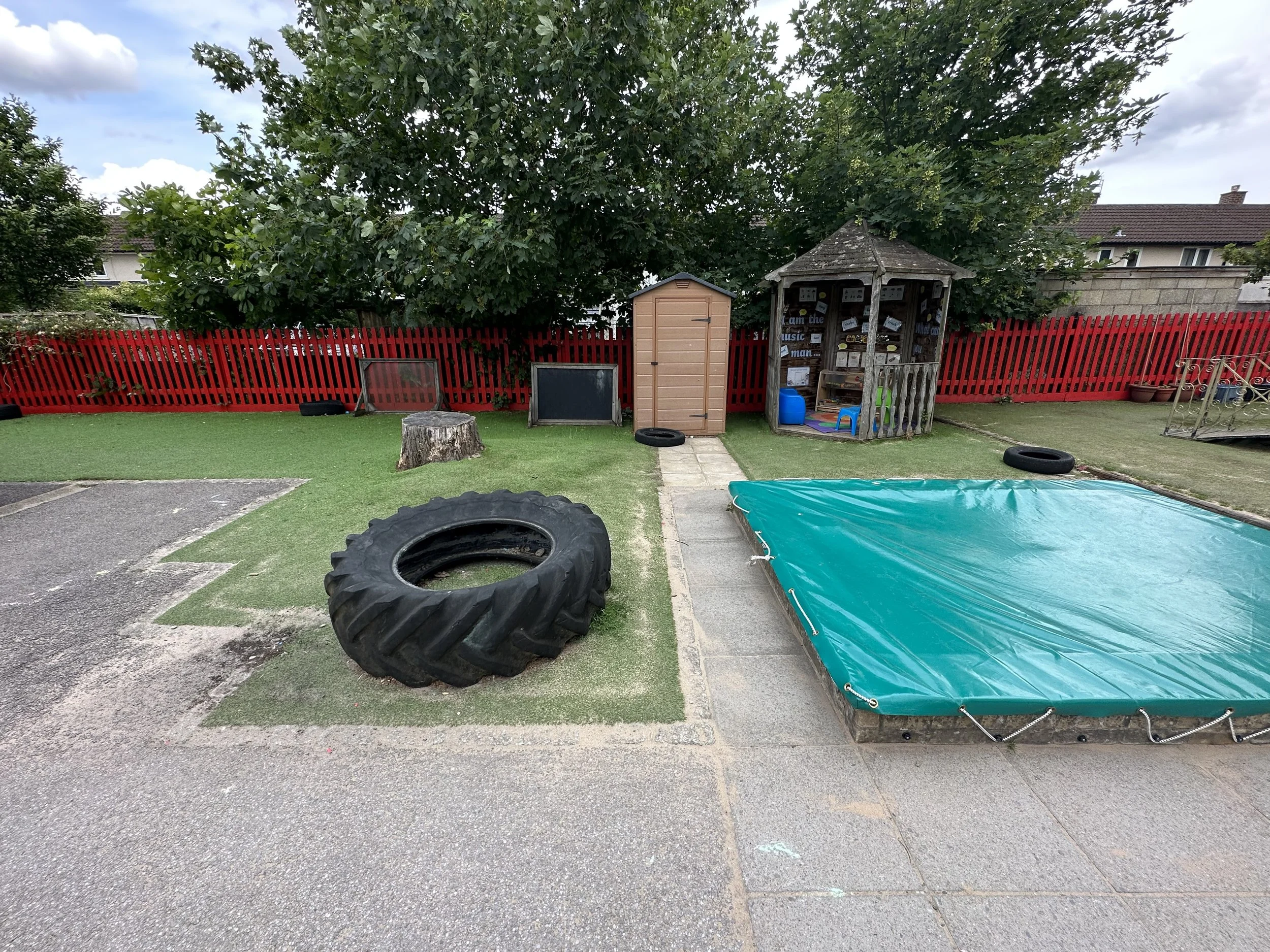A backyard playground area with a large tree, red fence, tire swings, a small shed, a wooden playhouse, a blue water container, a colorful mat, and various other toys and equipment.