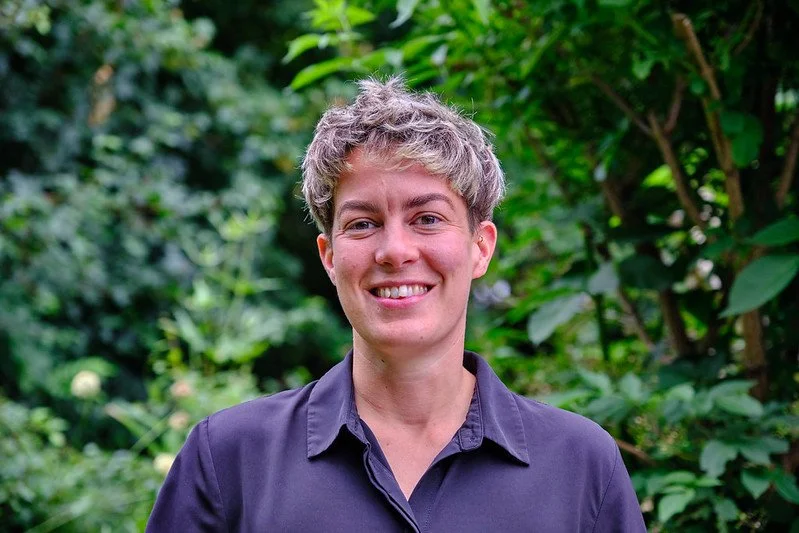 Young woman with short, curly blond hair smiling outdoors with green foliage in the background.