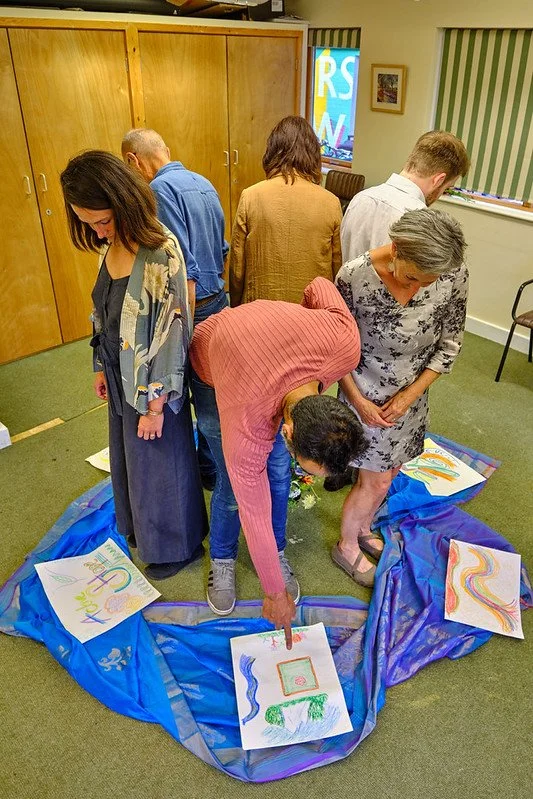 Group of six people gathered around colorful children's artwork on paper, placed on a blue fabric on the floor, in a room with wooden cabinets and a window.
